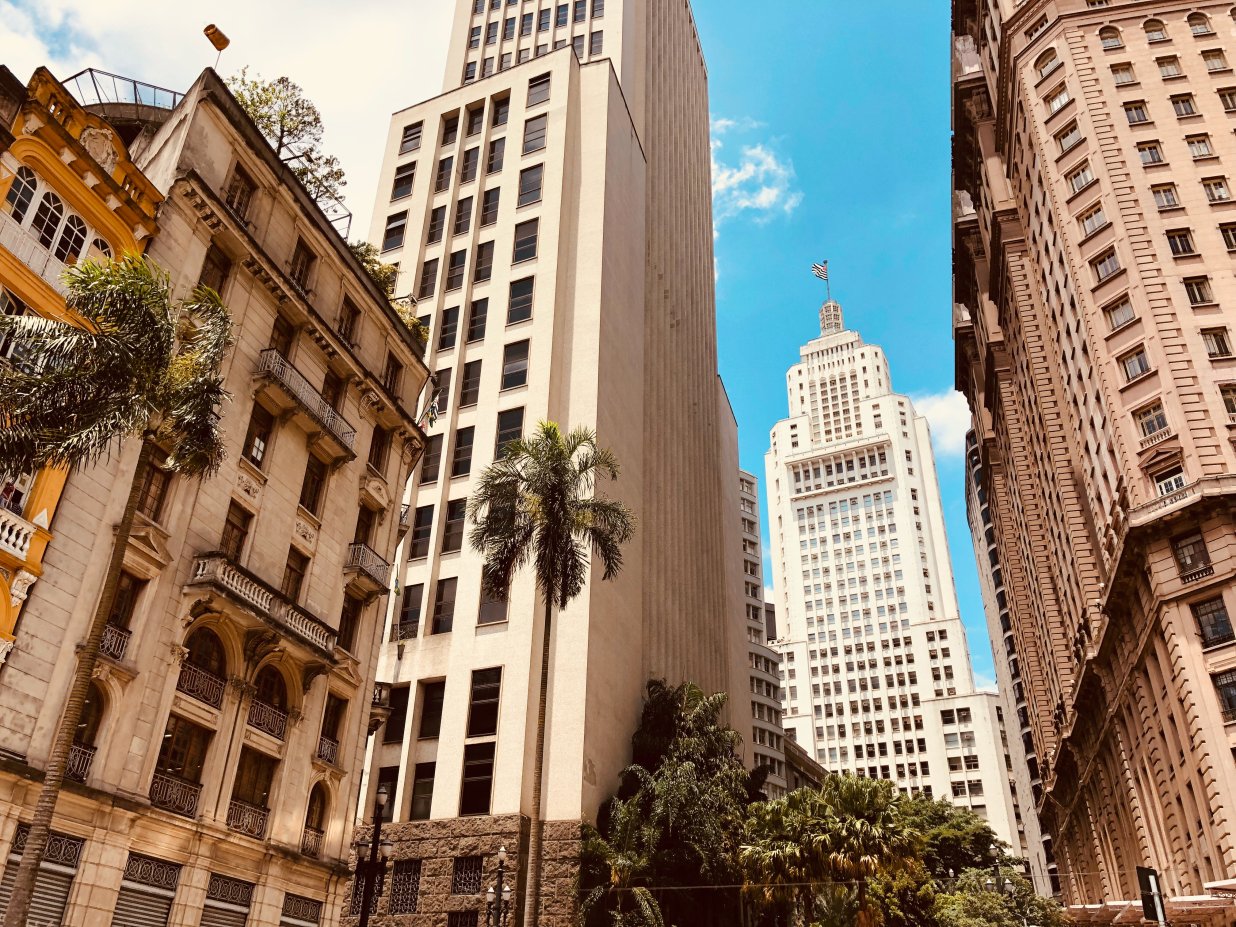 Auf dem Bild sieht man eine Stadtansicht von S&atilde;o Paulo, hoch aufragende Geb&auml;ude, davor Palmen an einem sonnigen Tag. | © © Joao Tzanno auf Unsplash