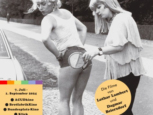 Lothar Lambert und Dagmar Beiersdorf 1979 bei den Dreharbeiten zu "Dirty Daughters" | © LoLa-Film/Dagmar Beiersdorf Auf der Straße stehend, hilft eine Frau einer aufreizend gekleideten Transfrau mittels eines Spiegels den Sitz ihrer sehr knappen Shorts zu überprüfen. | © LoLa-Film/Dagmar Beiersdorf