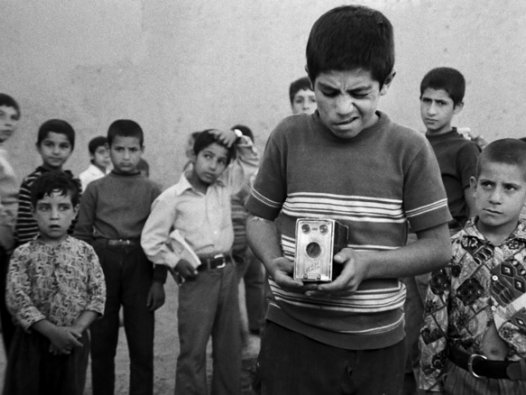 MOSSAFER / THE TRAVELLER | © Arsenal Auf dem Schwarz-Weiß-Foto sind mehrere Kinder zu sehen, im Vordergrund ein Junge, der einen Fotoapparat in den Händen hält und im Begriff ist, ein Foto zu schießen. | © Arsenal